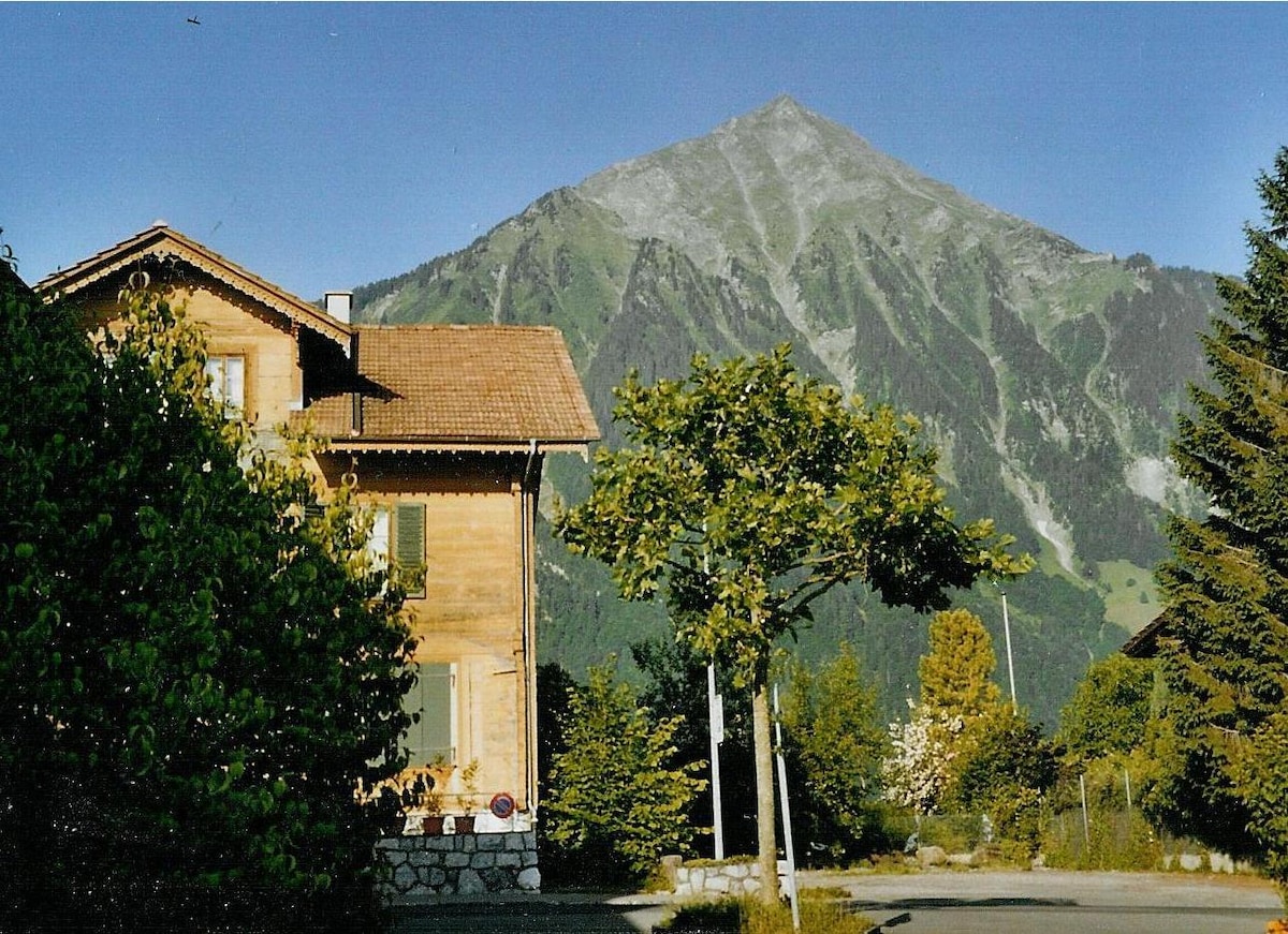 A traditional wooden house stands amidst lush greenery, framed by towering mountains in the background. The structure features a sloped roof and large windows, allowing natural light to filter inside. Trees and shrubbery surround the property, enhancing the serene outdoor environment.