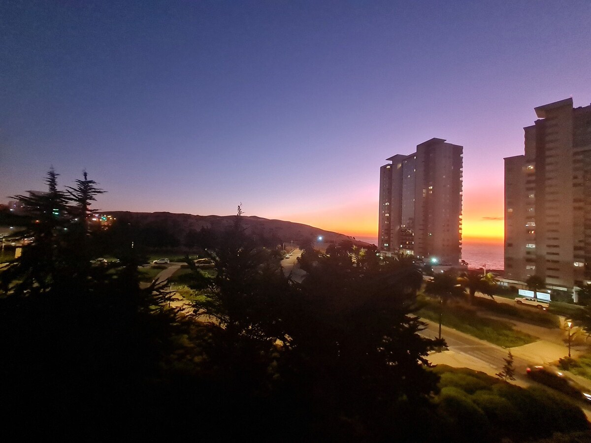 A serene twilight scene captures the skyline, showcasing tall buildings against a gradient of purple and orange hues as the sun sets. Palm trees and well-lit roadways frame the view, while the sea is visible in the distance.