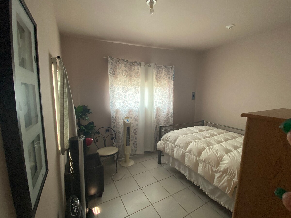 A cozy mini-studio apartment room is seen, featuring a spacious bed with a white duvet cover. Natural light filters through patterned curtains, illuminating the room. A tall mirror reflects the surroundings, while a small fan and a table add functionality to the space.