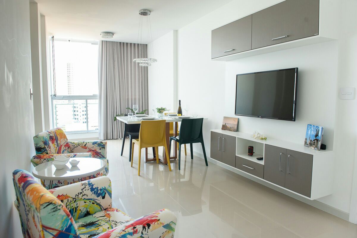 A bright living area features a large television mounted on a sleek cabinet. Colorful armchairs with floral patterns are positioned around a small coffee table, while a dining table for four sits nearby, illuminated by natural light from large windows.