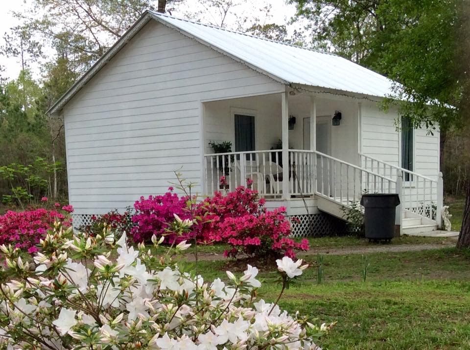 The cottage's exterior features a white facade with a metal roof, surrounded by vibrant azalea flowers in pink and white. A covered porch is visible, complete with chairs, while a walkway provides access to the entrance.