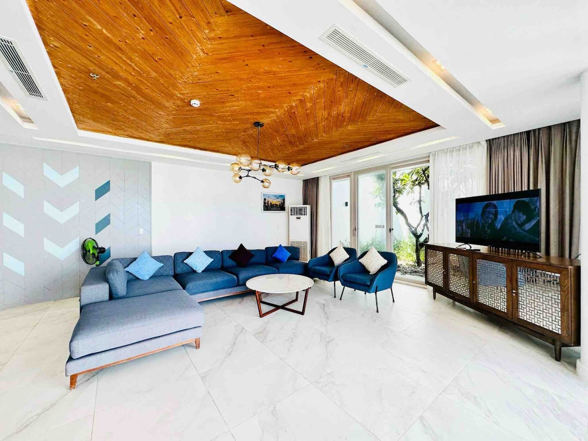 A spacious living area is brightened by natural light from large glass doors leading to an outdoor space. Comfortable seating is arranged around a circular coffee table, with blue accent pillows complementing the modern decor. A mounted television is positioned against a feature wall.
