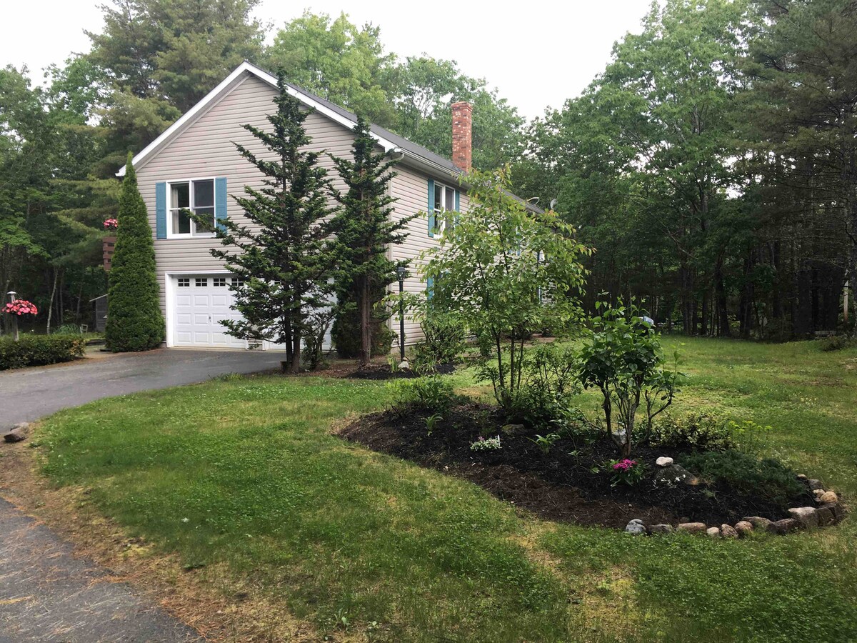 A two-story home is surrounded by a landscaped yard, featuring a mix of trees and shrubs. A manicured garden beds in front display various greenery and flowering plants. The driveway leads to a white garage door, offering ample space for parking.