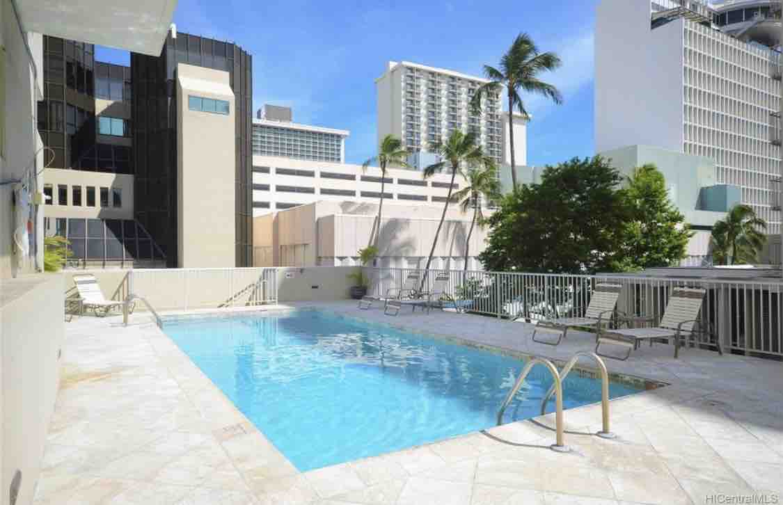 A tranquil pool area is displayed with crystal-clear water, surrounded by a marble deck. Several lounge chairs are positioned around the pool, offering a space for relaxation. Towering buildings and palm trees create a vibrant urban backdrop.