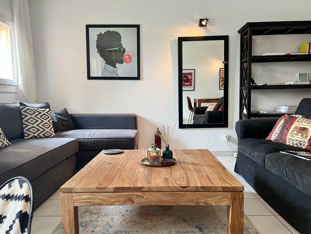 A cozy living area features a dark grey sectional sofa alongside a black armchair. A wooden coffee table with decorative items is centered in front, while a large mirror and an art piece adorn the wall. Natural light brightens the space through the window.