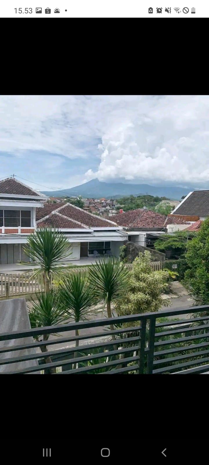 A scenic view from a balcony showcases traditional architecture with terracotta roofs and lush greenery. Surrounding palm trees add a tropical touch, while distant mountains are framed by clouds, providing a serene backdrop to the villas below.