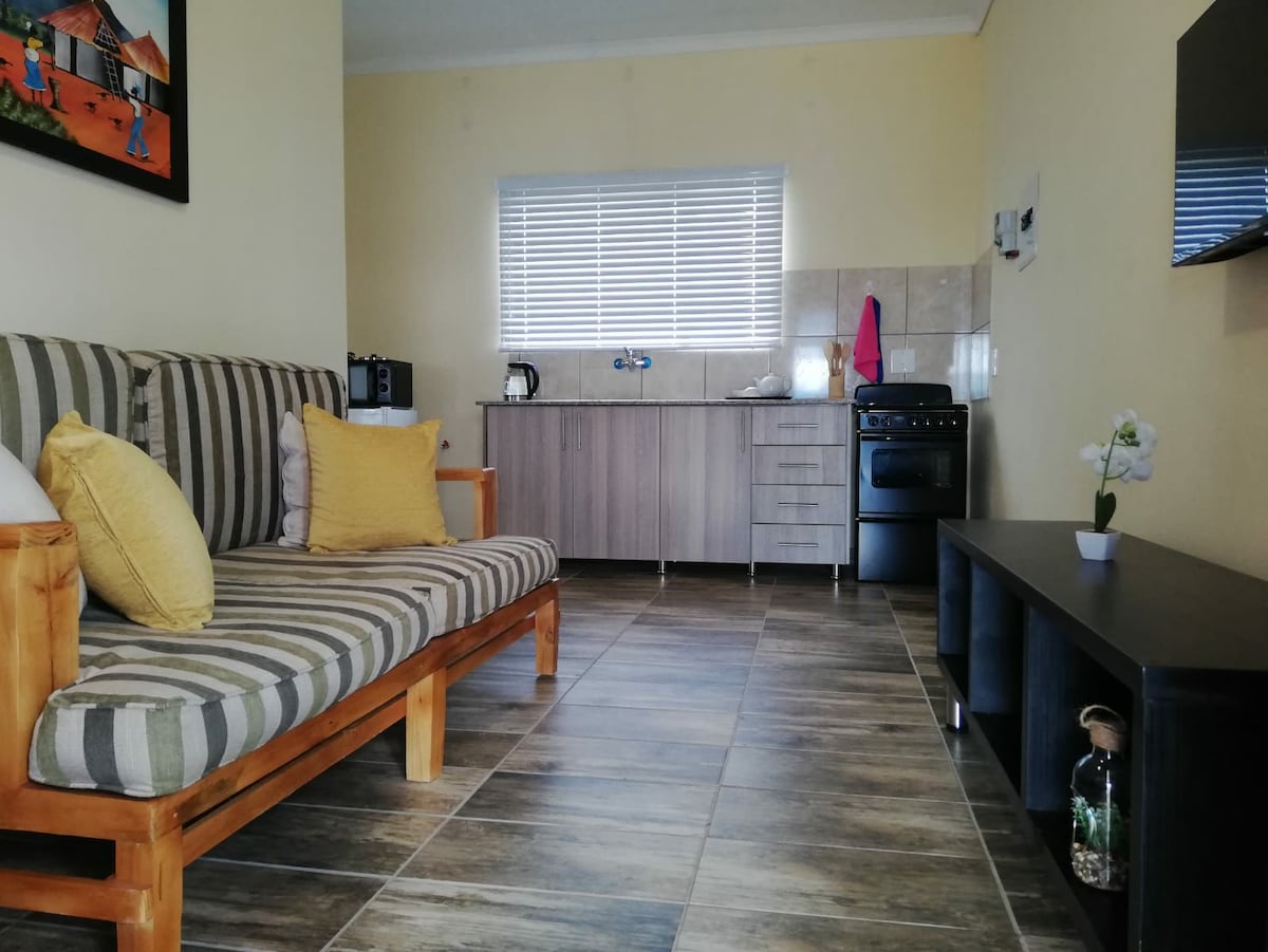 A cozy living area is displayed with a striped sofa and yellow cushions. To one side, a kitchen space features essential appliances, including a stovetop and microwave. Natural light enters through a window, and a TV is mounted on the opposite wall, providing a well-lit and functional space.