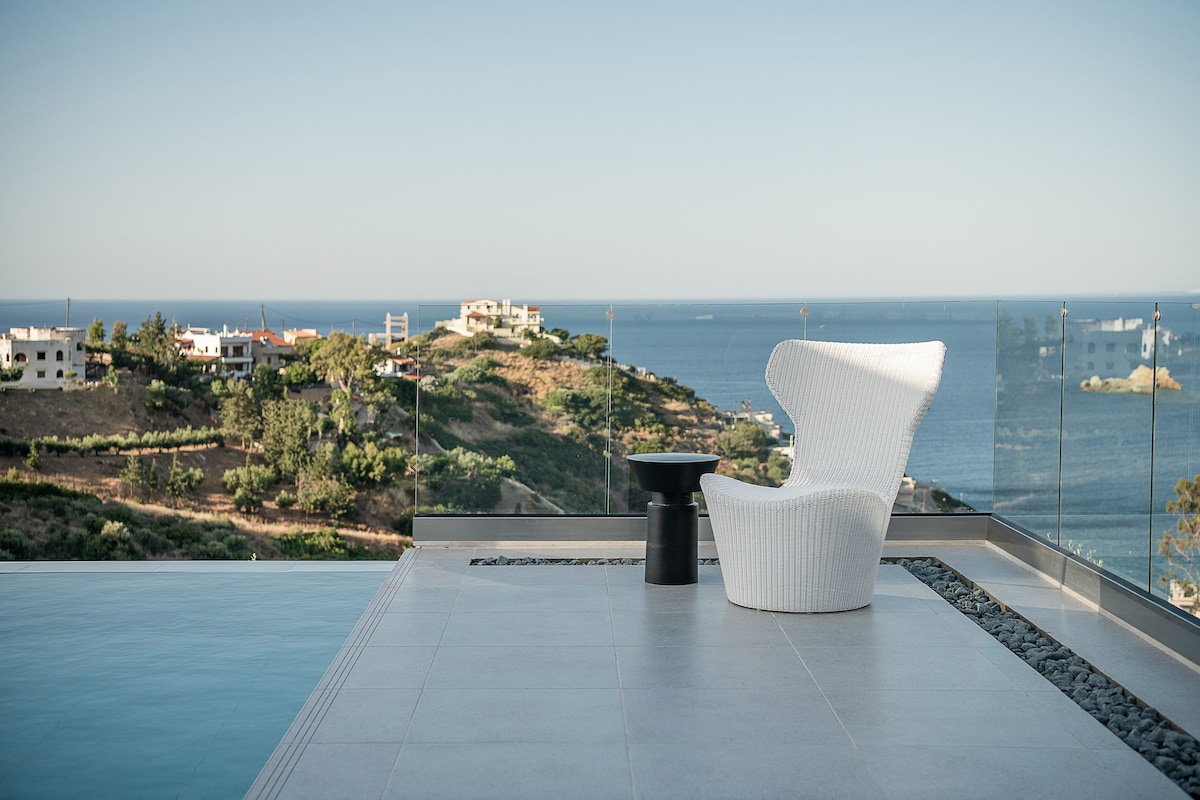 A white chair is positioned on a terrace overlooking panoramic views of the Cretan Sea and surrounding landscape. A small black table complements the seating area, and the adjoining infinity pool reflects the serene blue waters. The clear sky enhances the tranquil ambiance.