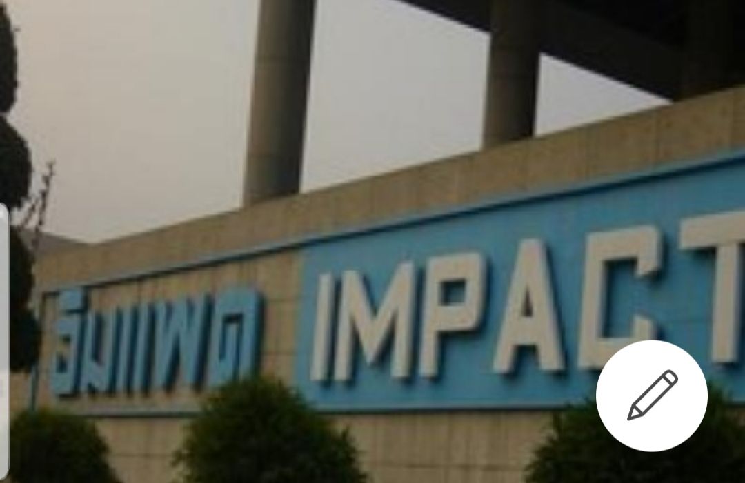 The image displays a large blue sign reading 'IMPACT,' prominently positioned in front of a concrete structure. Neatly trimmed shrubs are situated at the base of the sign, contributing to the clean lines of the area. The sky appears overcast, adding a muted tone to the scene.