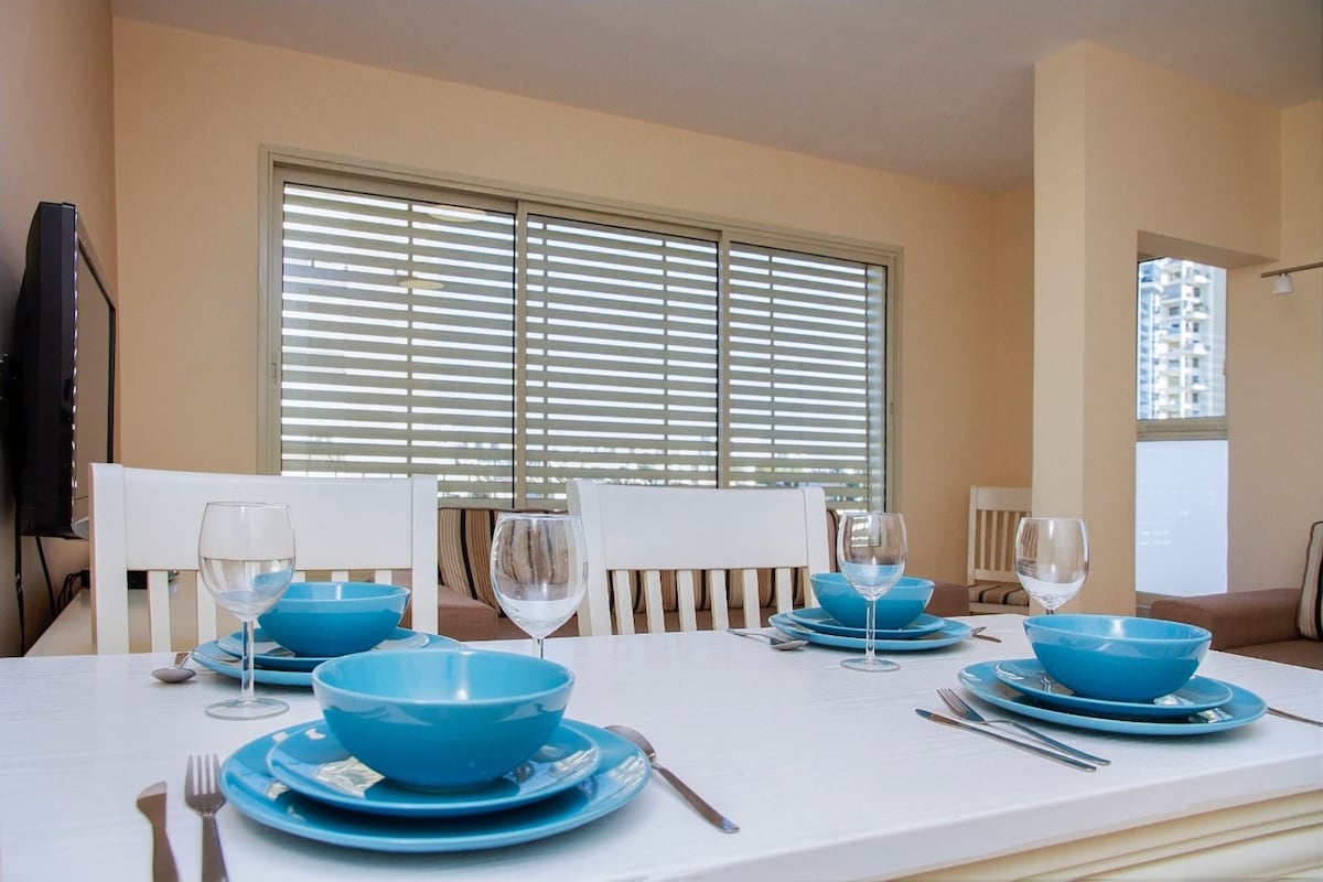 A dining area is presented with a large white table set for four. Blue plates and glassware rest on the table, while natural light filters through the stylish window coverings behind. A soft seating area can be glimpsed in the background.