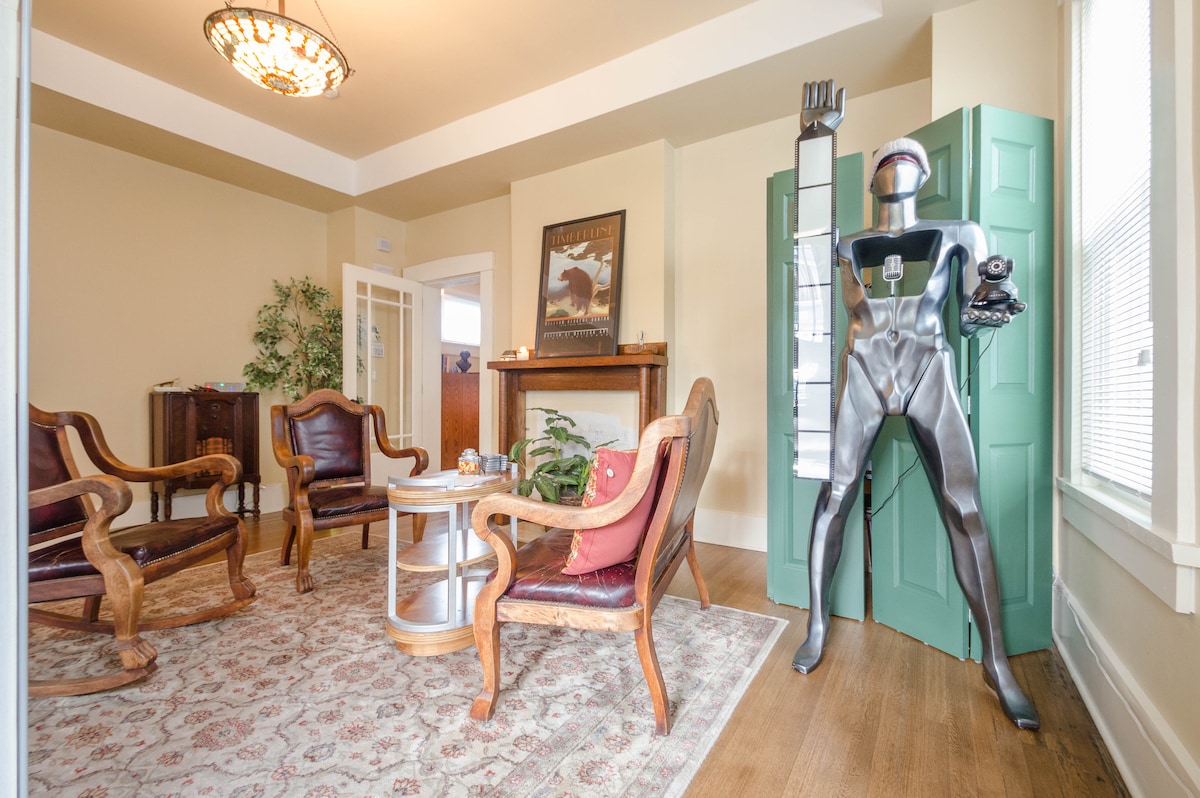 A welcoming sitting area features four wooden chairs with leather accents arranged around a round coffee table. A decorative area rug covers the wooden floor, and a surveillance bot stands in the corner next to a green door. Natural light filters in through the windows.