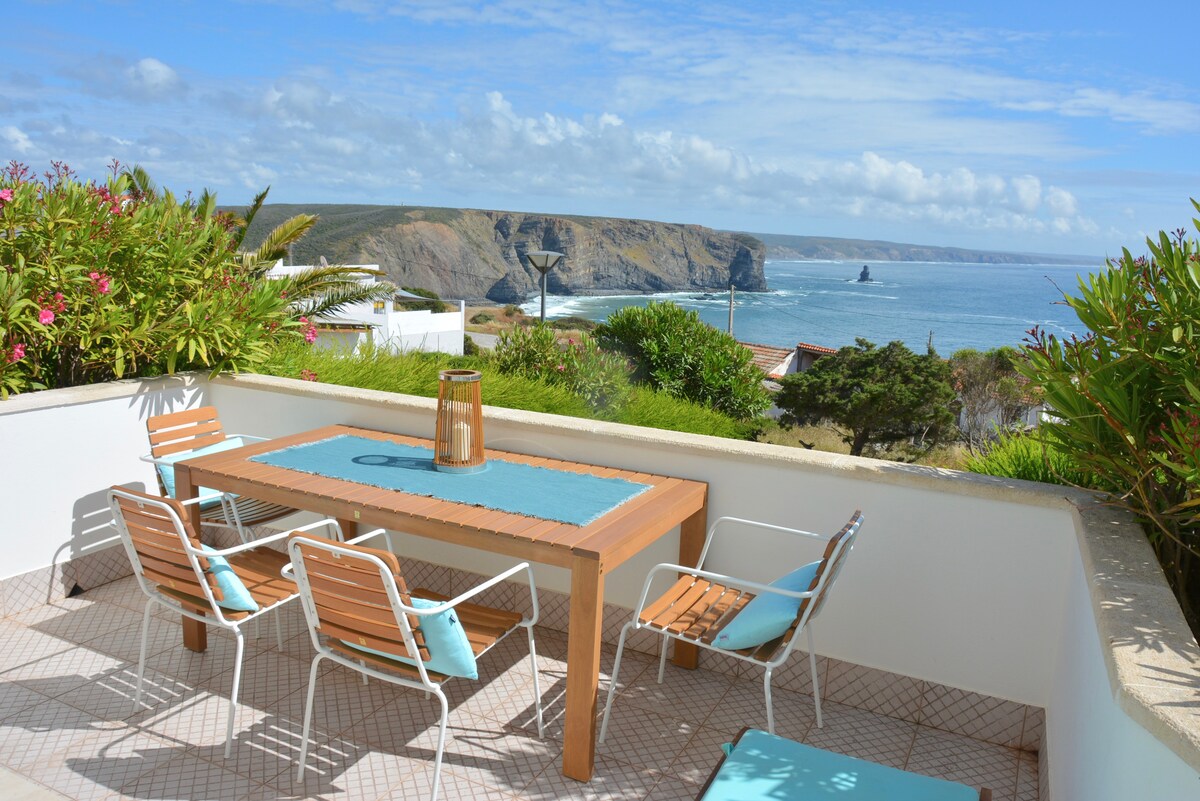 An outdoor terrace offers a wooden dining table surrounded by six chairs, providing a scenic view of the coastline and ocean. Lush greenery frames the space, with clear blue skies and gentle waves visible in the background, creating a serene setting.