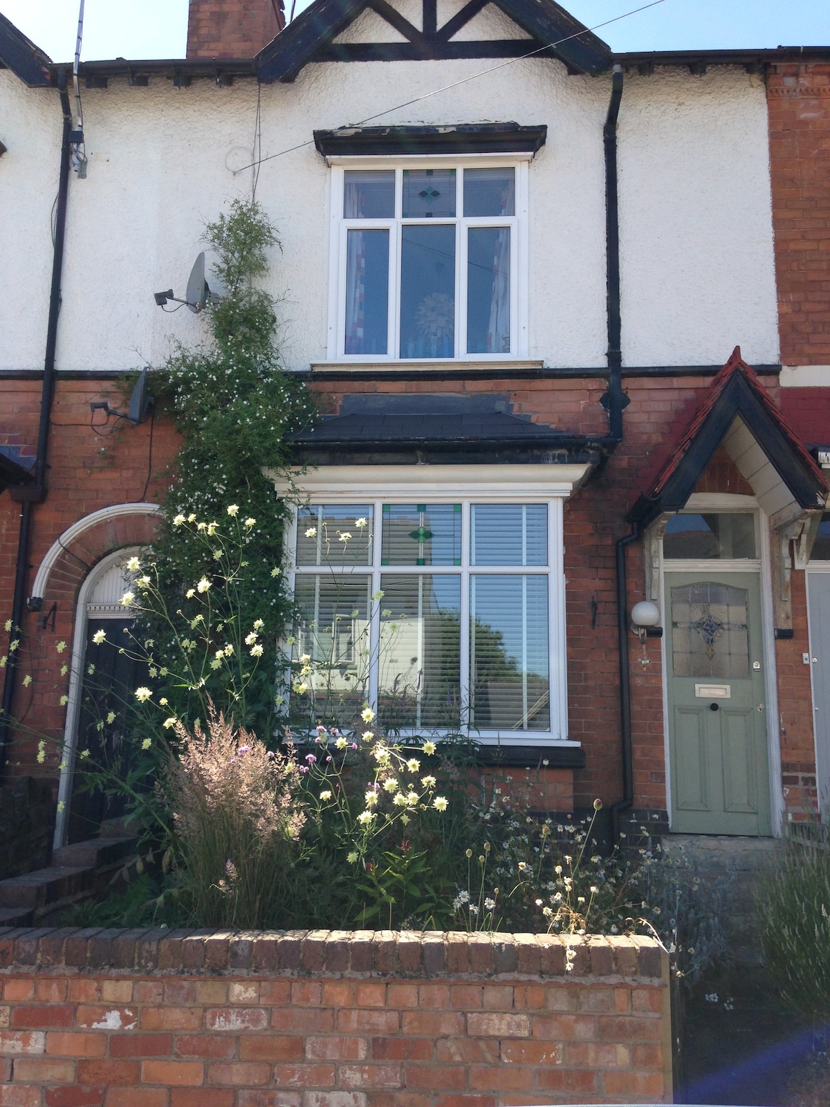 The exterior of a charming Edwardian house is shown, featuring a front garden with wildflowers and greenery. A large window allows natural light to fill the interior. Decorative elements including a second story window, satellite dish, and traditional brickwork enhance the character of the property.