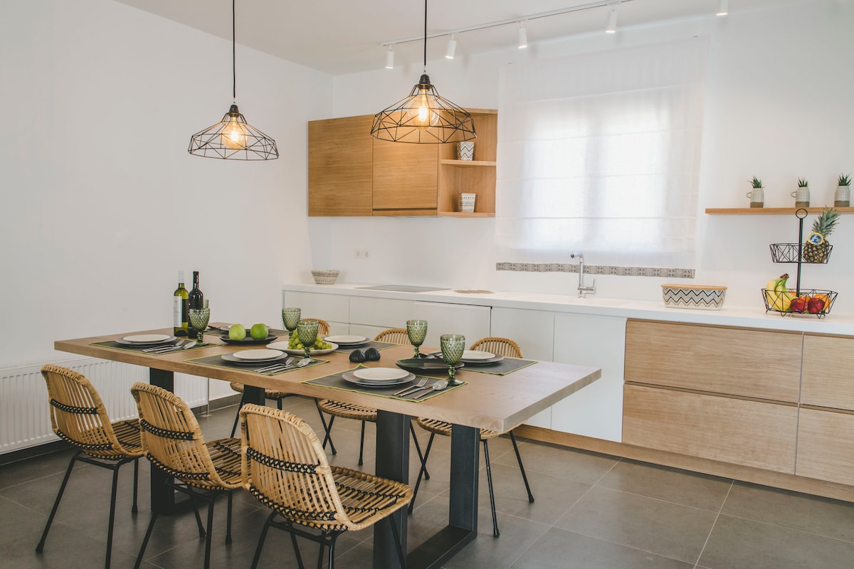A modern kitchen and dining area feature a wooden dining table with stylish chairs arranged around it. Elegant pendant lights hang above, while minimalistic cabinetry provides storage. A window allows natural light to brighten the space, enhancing the clean, contemporary design.