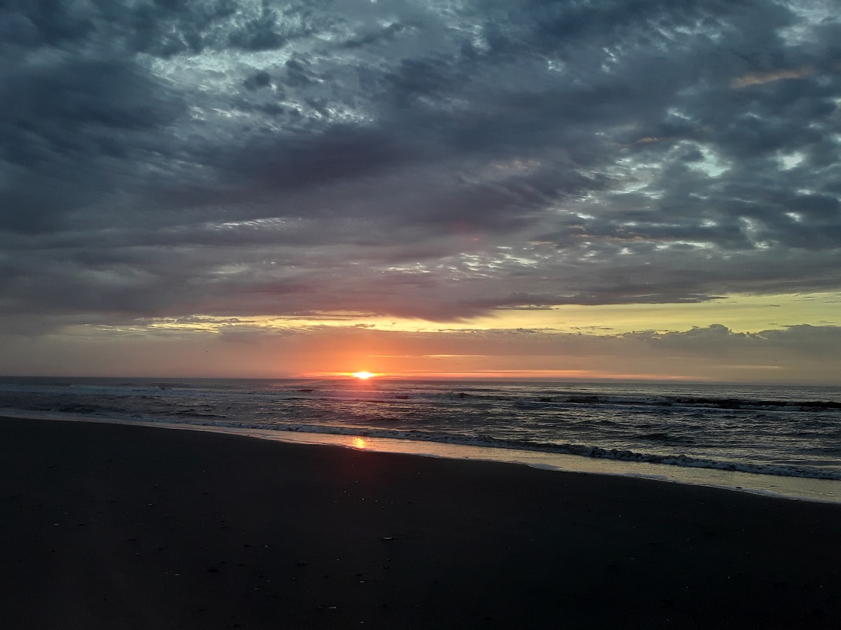 The image captures a serene beach scene during sunset. Soft, warm colors fill the sky as the sun sets on the horizon, reflecting gently on the water. Calm waves create a rhythmic pattern along the shore, enhancing the peaceful ambiance.