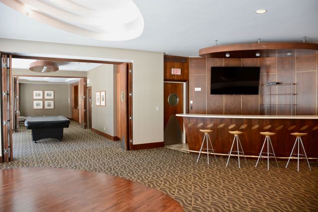 A spacious party room is shown, featuring a sleek bar with high stools, a large television, and a pool table in the background. The soft, neutral tones of the walls and floor complement the warm wood accents of the bar area.