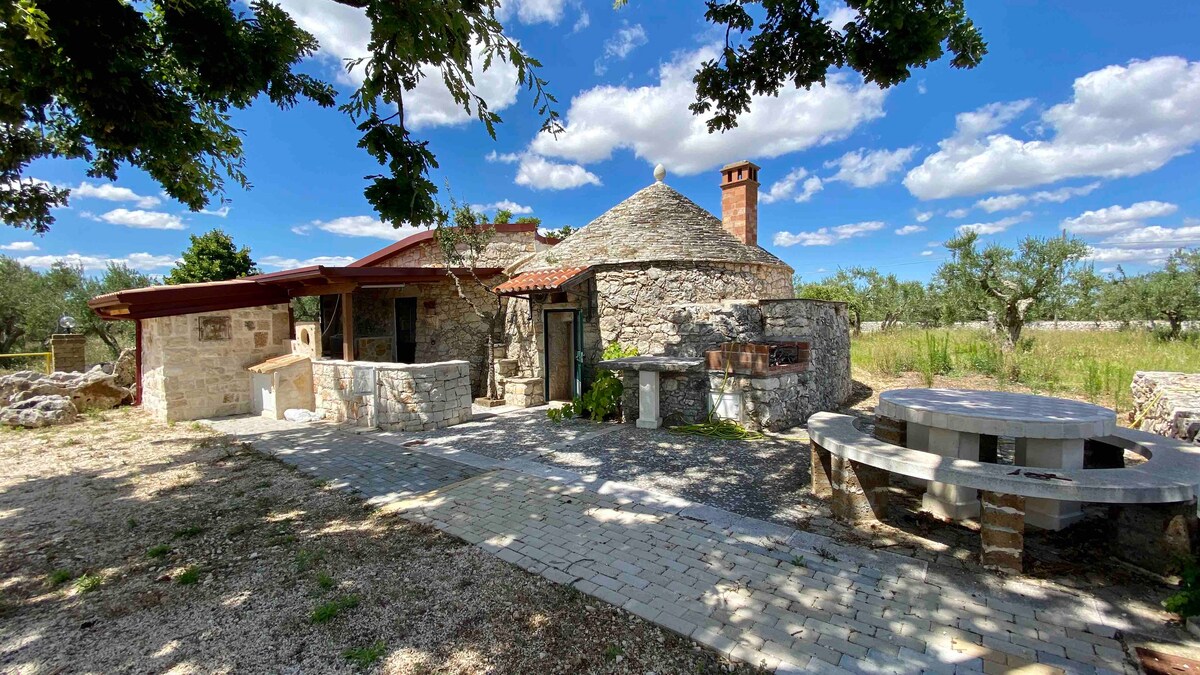 A rustic stone structure with a red-tiled roof is surrounded by greenery, set against a bright sky with fluffy clouds. A spacious outdoor area features a stone barbeque and a round seating arrangement made from stone. Sunlight creates soft shadows across the pathway.