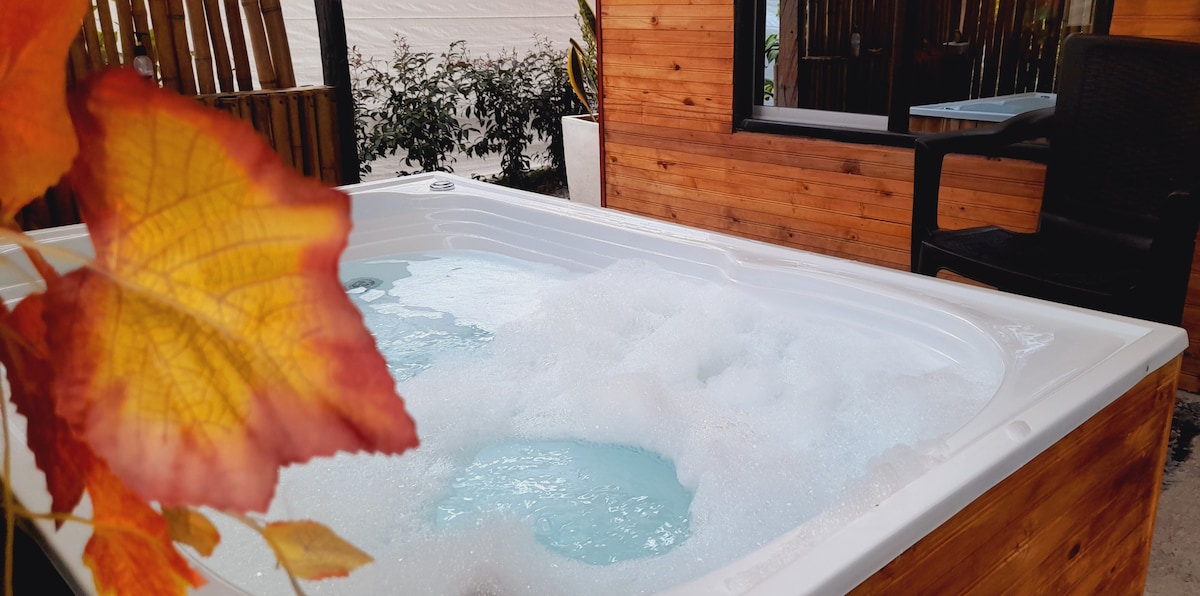 A jacuzzi filled with bubbles showcases a tranquil blue hue, surrounded by wooden paneling. A black chair is positioned nearby, and foliage can be seen in the background, adding a touch of natural color to the serene setting.
