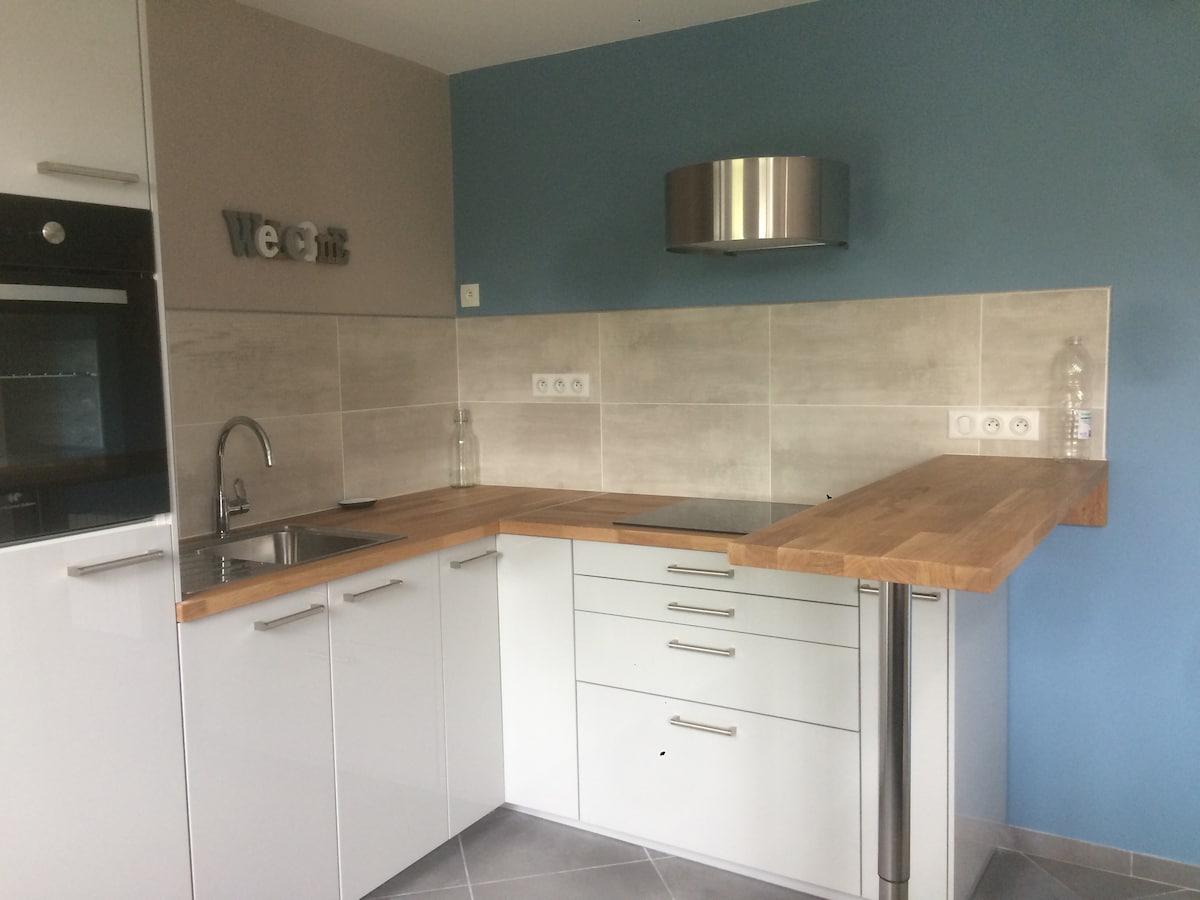 A modern kitchen area features a sleek countertop made of wood, complemented by white cabinetry. The walls are painted in a calming blue and light gray tones. A stainless steel hood is installed above the stove, with essential appliances neatly arranged for convenience.