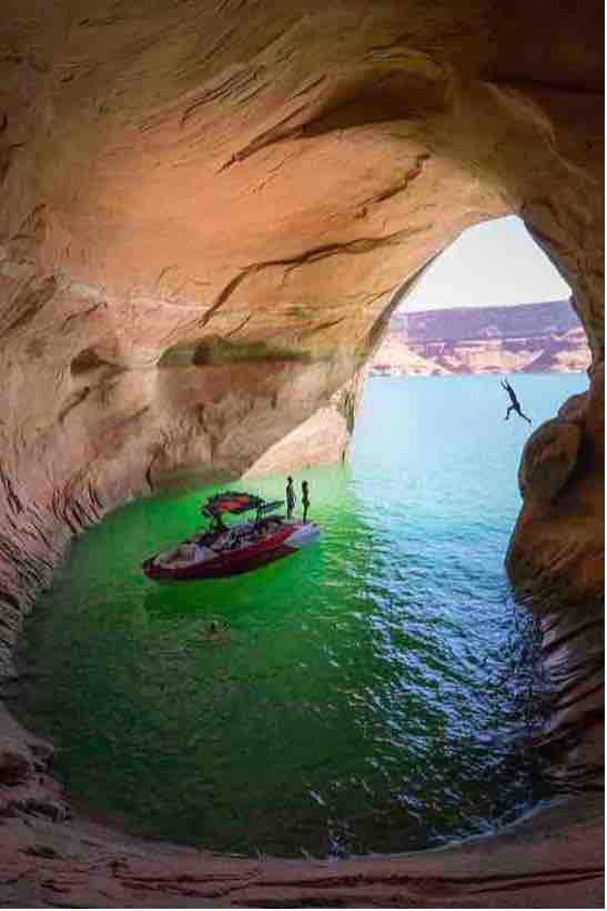 A natural rock formation creates an opening that frames a serene body of green water. A boat is anchored nearby, while individuals are seen engaging in activities. The cave's smooth, warm-toned walls contrast with the cool hues of the water.