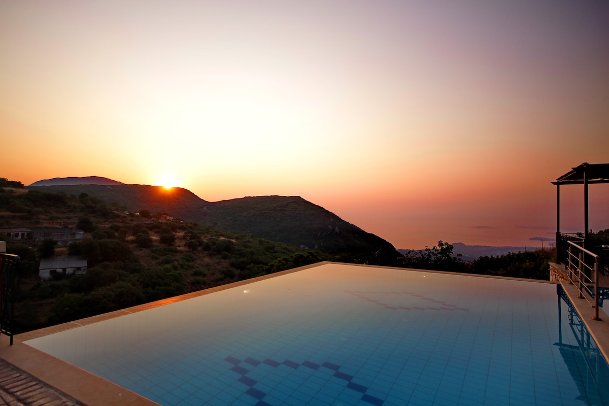 A serene infinity pool reflects soft hues of orange and pink at sunset, with hillsides gently rolling in the background. The tranquil scene is accentuated by the vast sky, creating a peaceful ambiance that invites relaxation.
