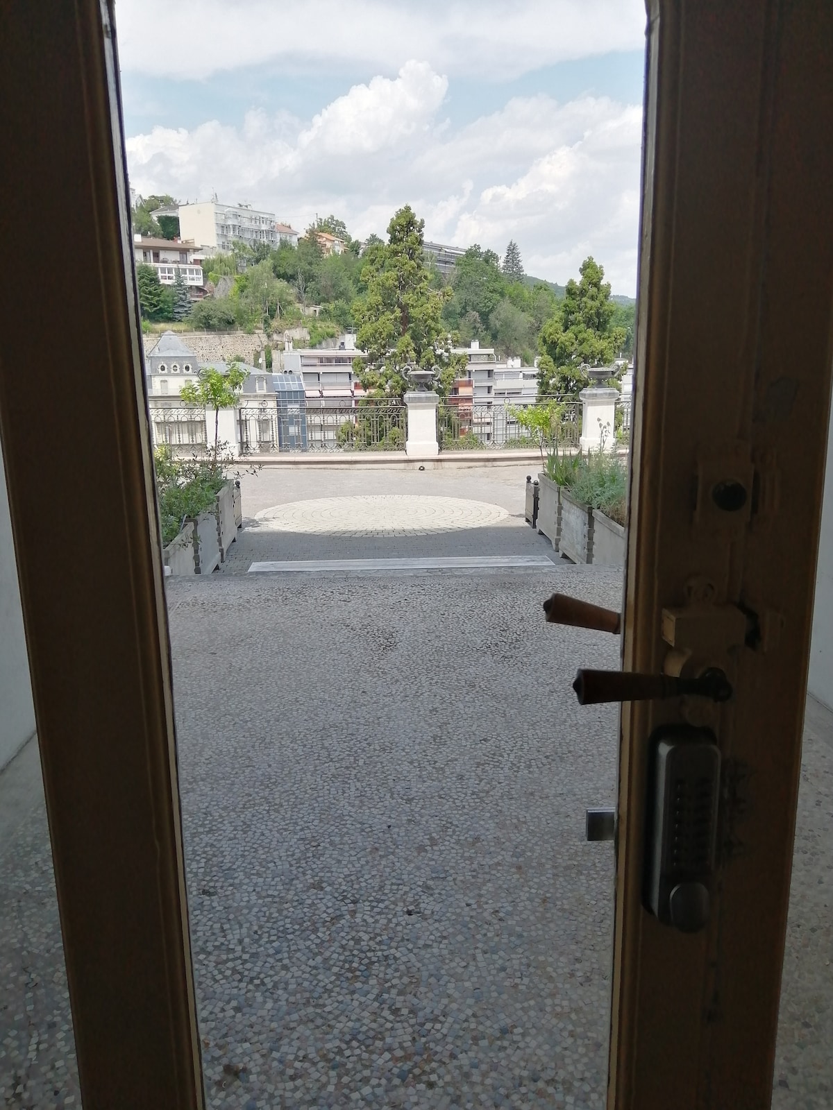 An outdoor view is visible through a partially opened door, showcasing a pebbled path leading to a landscaped area. Green trees line the background, and residential buildings can be seen atop the hills. The sky is partly cloudy, inviting a calm atmosphere.
