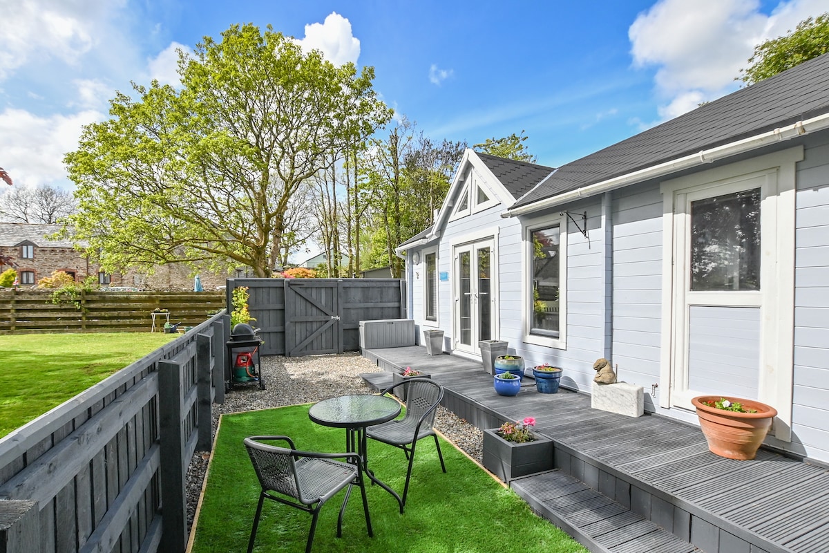 A private garden area is depicted featuring a small table with two chairs on a grassy surface. Potted plants with colorful blooms are placed around the space. The studio is shown to have large windows, allowing natural light to brighten the exterior.