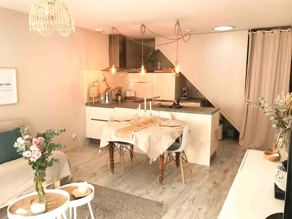 A modern and cozy interior features a dining area with two chairs and a table set for a meal. Warm pendant lights hang above the kitchen counter, where appliances and decor are visible. A seating area includes neutral-toned furnishings and a floral arrangement.