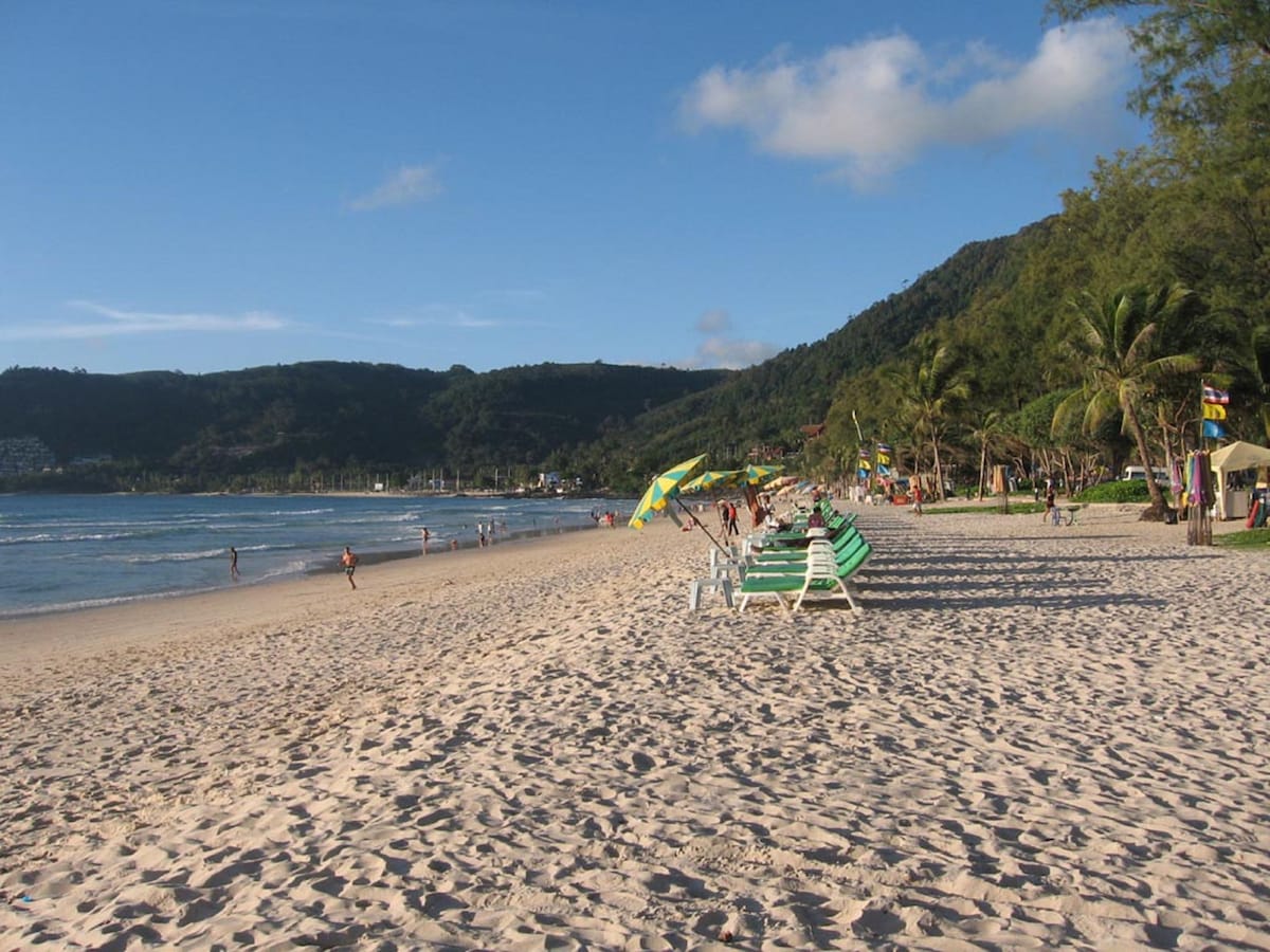 A sandy beach stretches along the coastline, with gentle waves lapping at the shore. Green and white striped loungers are set up on the sand, while palm trees gently sway in the background. Beachgoers can be seen enjoying the sun and the scenic mountain views.