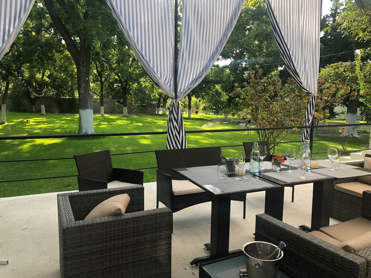 An outdoor seating area features a set of dark wicker chairs and tables, arranged under elegant striped canopies. Lush green grass and trees provide a serene backdrop, creating a space for relaxation and connection with nature.