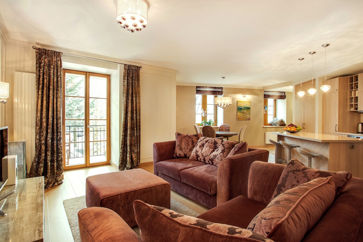 A spacious living room features plush sofas adorned with patterned throw pillows. Natural light filters through the large windows, illuminating the warm, neutral tones of the decor. A modern dining area is visible in the background, complemented by pendant lighting and wood accents.