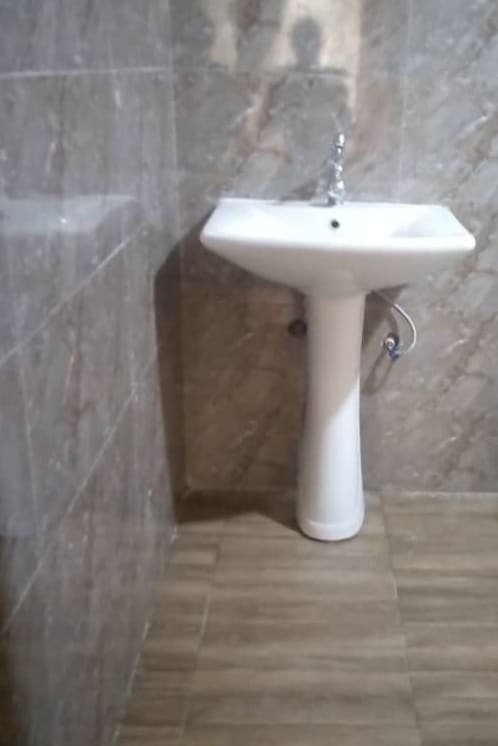 A clean bathroom is showcased with tiled walls and a modern pedestal sink. The flooring features wood-like tiles, creating a warm atmosphere. The space is well-maintained, emphasizing simplicity and functionality.