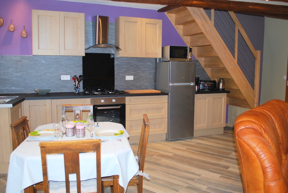The kitchen area features modern wooden cabinetry and a sleek countertop, equipped with essential appliances including a refrigerator and stove. A dining table with four wooden chairs is set for a meal, complemented by a staircase leading to the upper level.