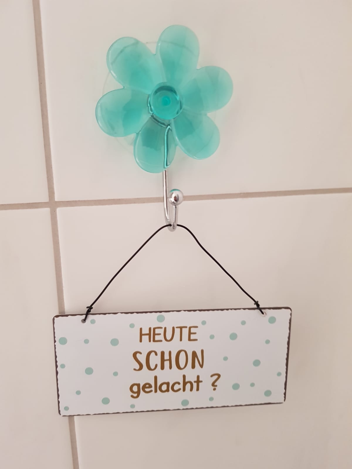 A teal flower-shaped hook is mounted on the wall, supporting a decorative sign. The sign, featuring a white background with teal polka dots, contains the text 'HEUTE SCHON gelacht?' in gold lettering, inviting lightheartedness in the space.