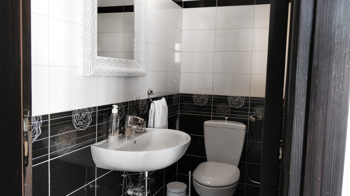 A modern bathroom is displayed, featuring white and black tiled walls. A round sink with a chrome faucet is positioned next to a toilet. A rectangular mirror with an ornate frame is mounted above the sink. Towels and toiletries are neatly arranged.