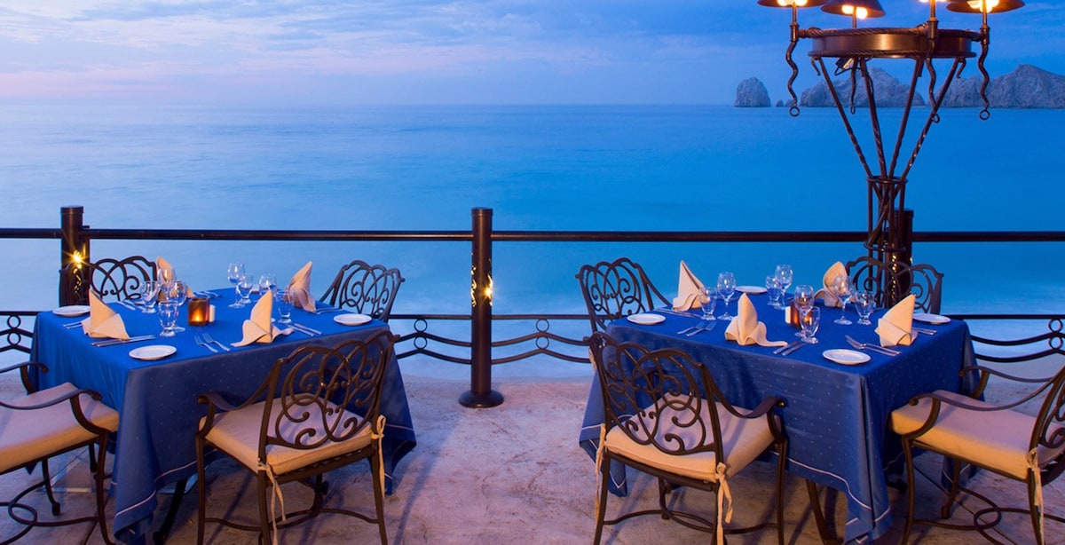 A dining area is set for an outdoor meal, featuring round tables draped with blue tablecloths and elegant white napkins. A stunning ocean view is visible in the background, with the horizon meeting the sky, enhancing the tranquil ambiance of the space.