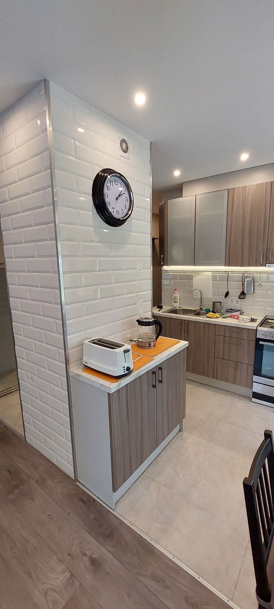 A modern kitchen features sleek cabinetry with wooden finishes and a glossy white backsplash. Equipped with essential appliances, a counter toaster sits next to a coffee maker. Bright overhead lights enhance the clean, organized layout, complemented by a wall clock displayed prominently.