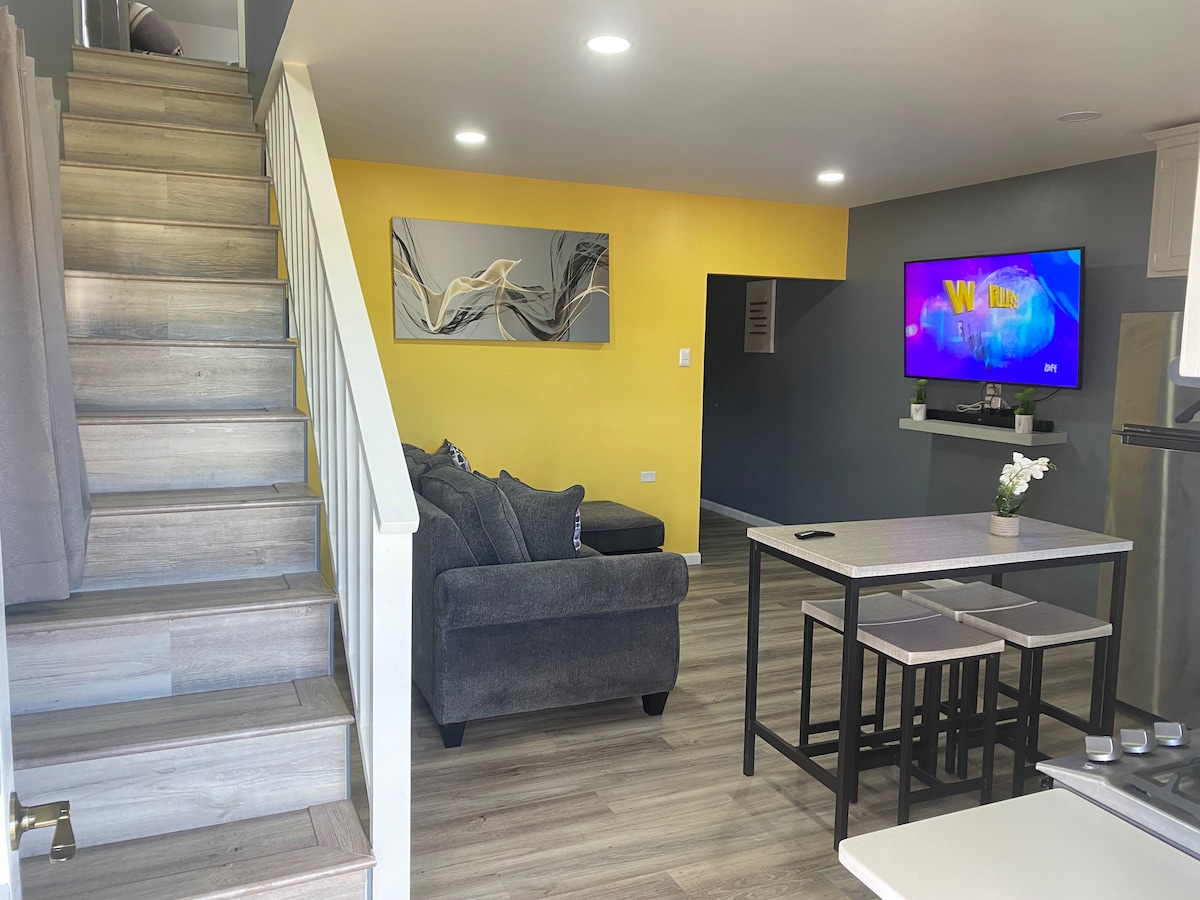A well-lit living area features a staircase leading to the upper level. A dark gray sectional sofa is positioned near a wall-mounted television, which displays colorful content. A dining table with four chairs is situated in the foreground, with a decorative vase on the table.