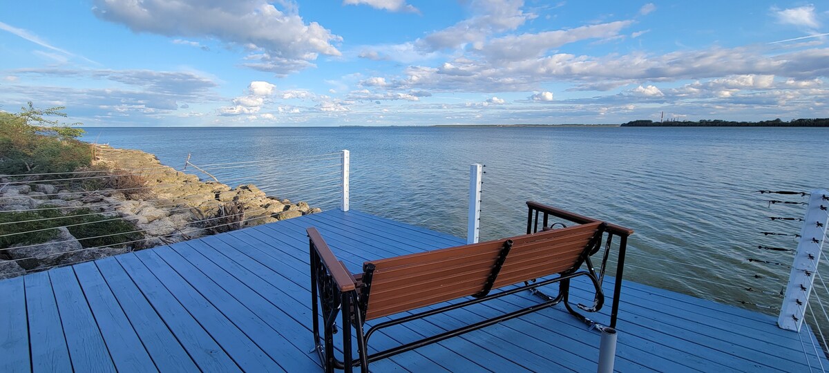 A wooden bench is positioned on a bright blue deck overlooking the calm waters of Lake Erie. The scene is framed by clear skies dotted with fluffy clouds, while rocky shorelines provide a natural border.