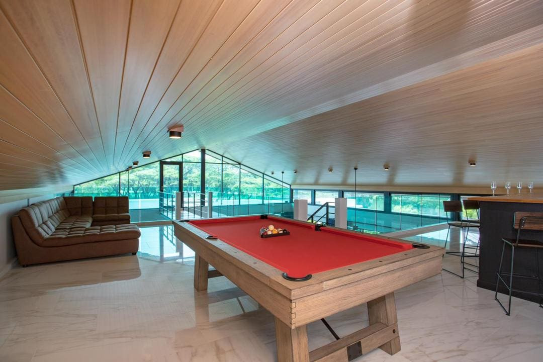 An upper-level game area features a pool table with red felt, surrounded by sleek tile flooring. A comfortable sofa is positioned nearby, offering a space for relaxation. Large glass windows provide expansive views of the surrounding greenery, enhancing the open and airy atmosphere.
