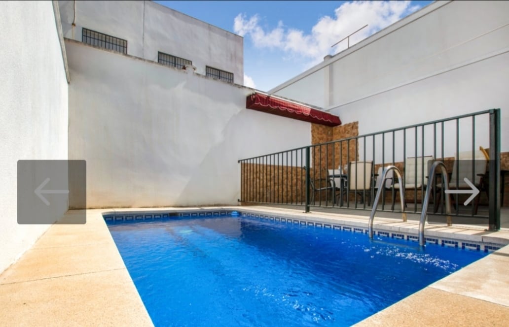 A private pool is surrounded by a low fence, offering a safe space for relaxation. Sunlight reflects off the clear blue water, while a small dining area with a table and chairs is present nearby, set against a white wall under a shaded awning.