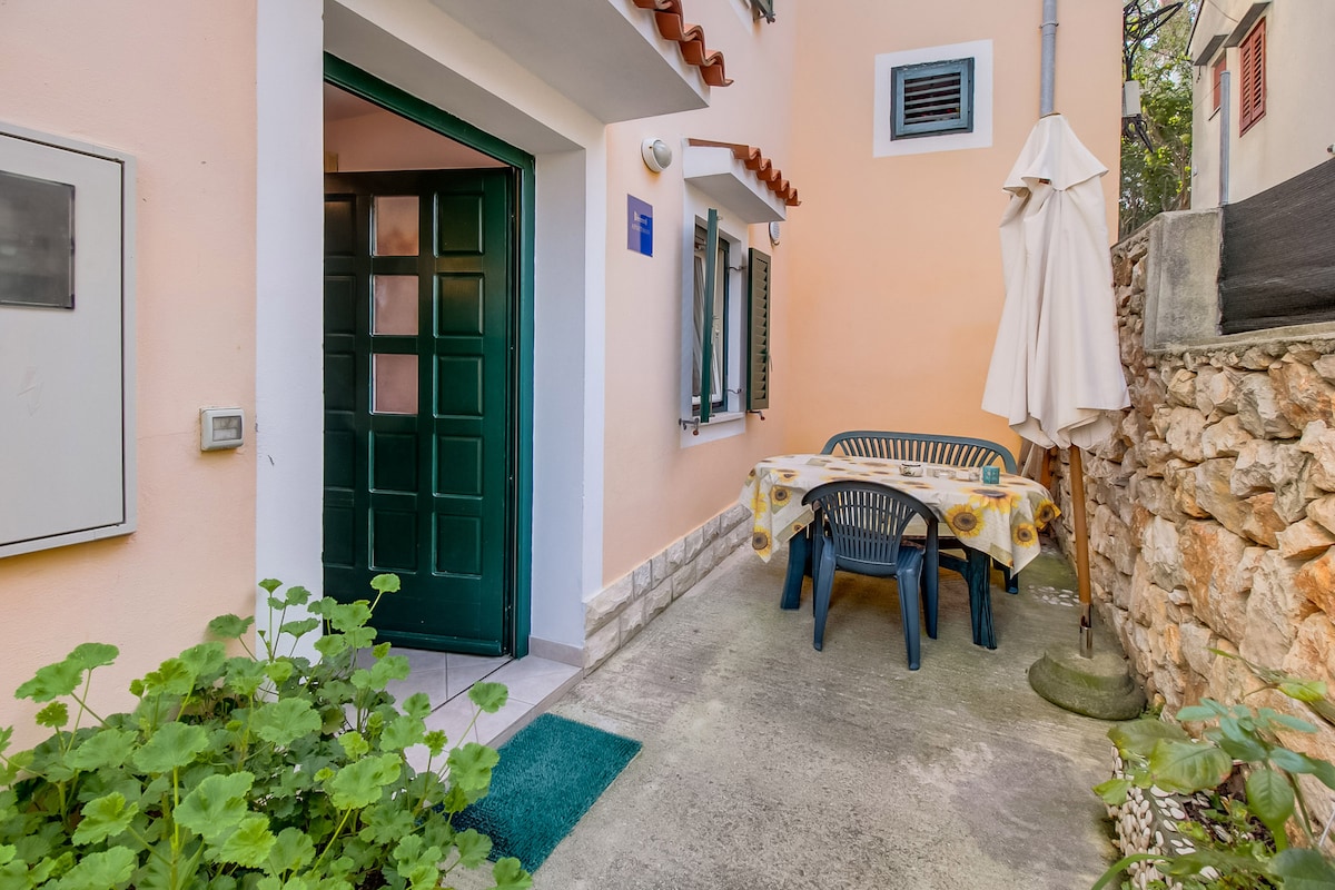 An outdoor terrace features a dining table surrounded by four dark plastic chairs. The entrance is framed by a green door, and an umbrella provides shade. The surrounding area includes potted plants and a stone wall, contributing to a serene and private setting.