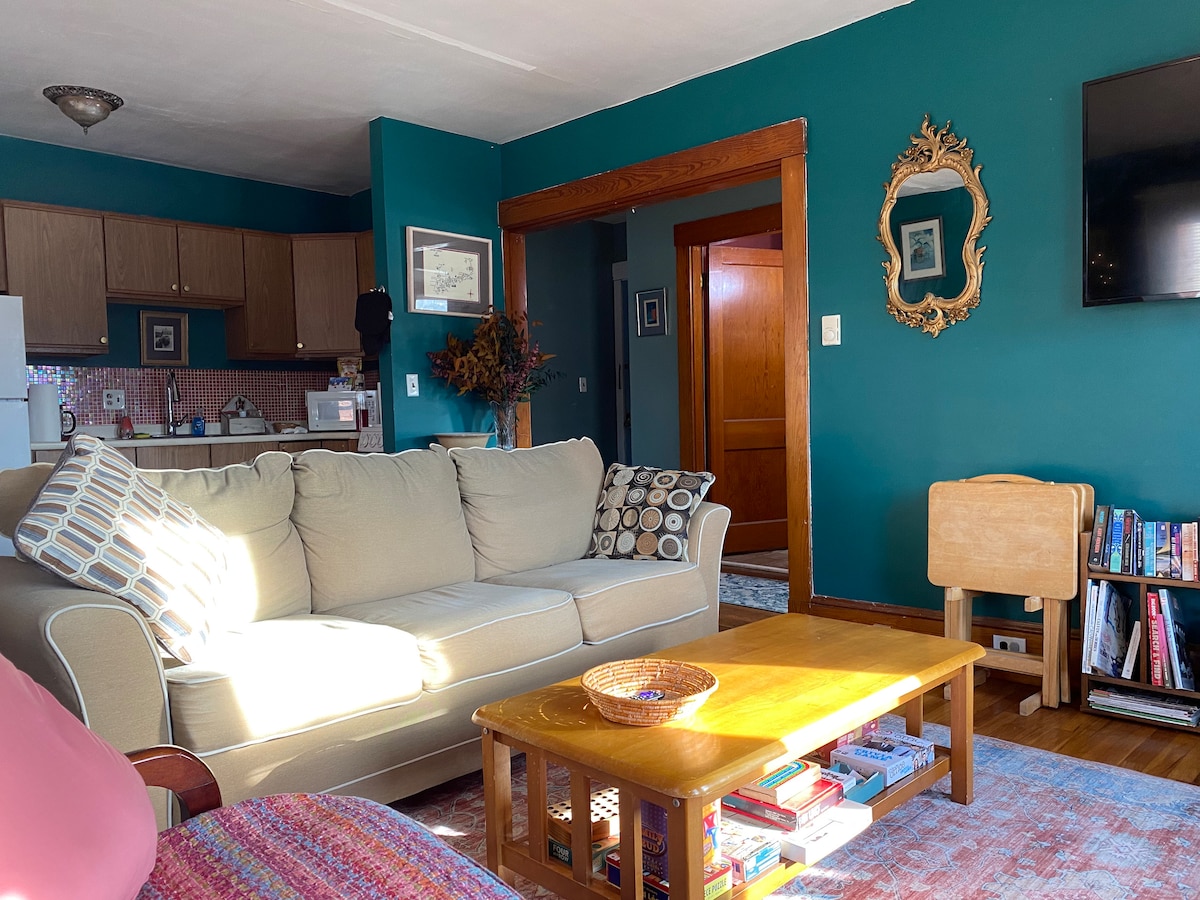 A cozy living area is highlighted by a light-colored sofa adorned with decorative cushions. Wooden furniture, including a coffee table and bookshelf, add warmth. Bright walls in teal contrast with the subtler tones of the kitchen area seen in the background.