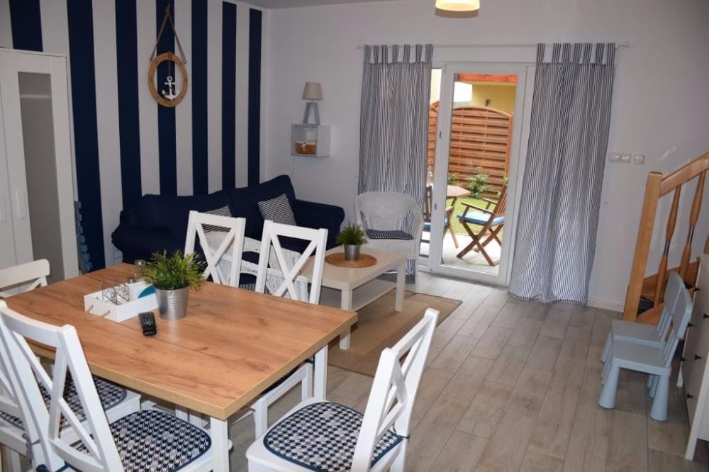 A bright living area features navy and white striped walls, a wooden dining table surrounded by six chairs, and a cozy sofa. Natural light flows through glass doors leading to a private terrace. Decorative plants add a fresh touch to the space.