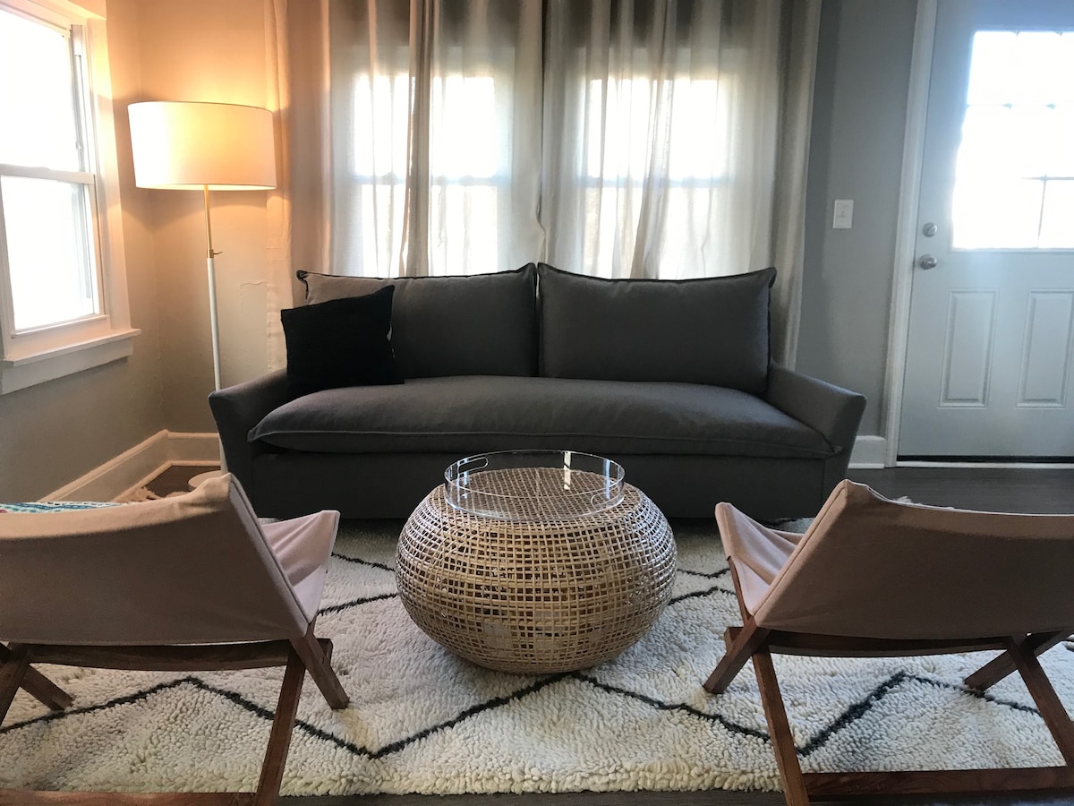 A cozy living area features a gray pullout sofa, two light-colored armchairs, and a round woven coffee table. Natural light fills the space through large windows, which are dressed with sheer curtains. A neutral area rug adds warmth to the floor.