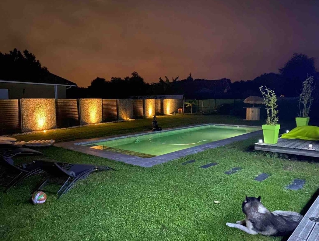 An inviting outdoor space is showcased, featuring a well-lit pool surrounded by lush grass. Lounge chairs are positioned nearby, and a decorative plant adds a touch of greenery. A calm evening atmosphere is complemented by soft lighting along the perimeter.