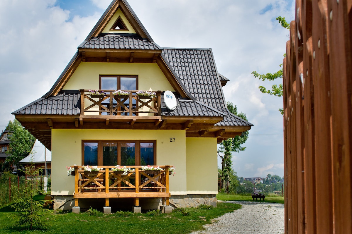 A charming house with a sloped roof and wooden balcony is nestled among greenery. Large windows allow natural light to fill the interior. The front porch features flower boxes, enhancing the home’s inviting exterior. A gravel path leads to the entrance, surrounded by a peaceful landscape.