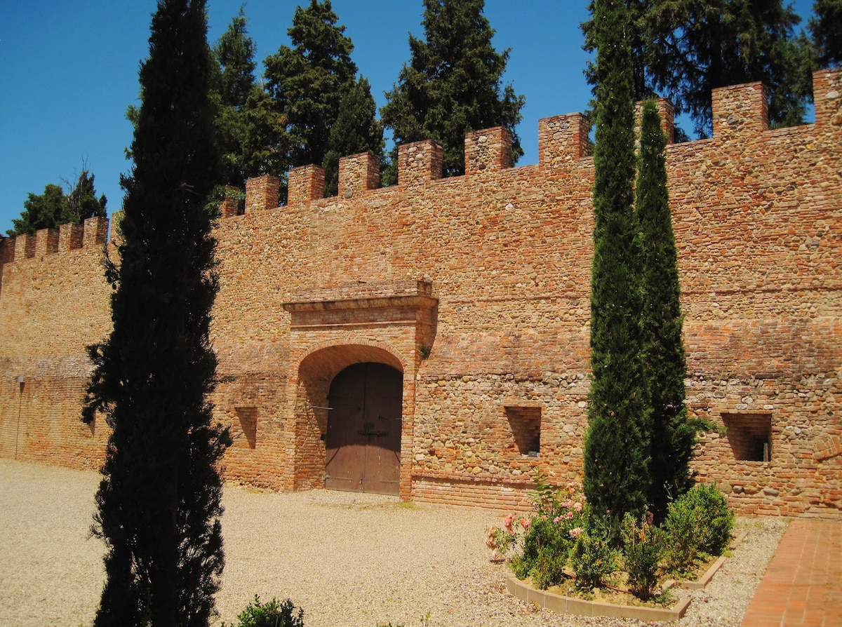 A historic stone wall surrounds a courtyard, featuring a large arched entrance with wooden doors. Tall cypress trees flank the entrance, and lush greenery can be seen at the base of the wall. The bright blue sky provides a contrasting backdrop.