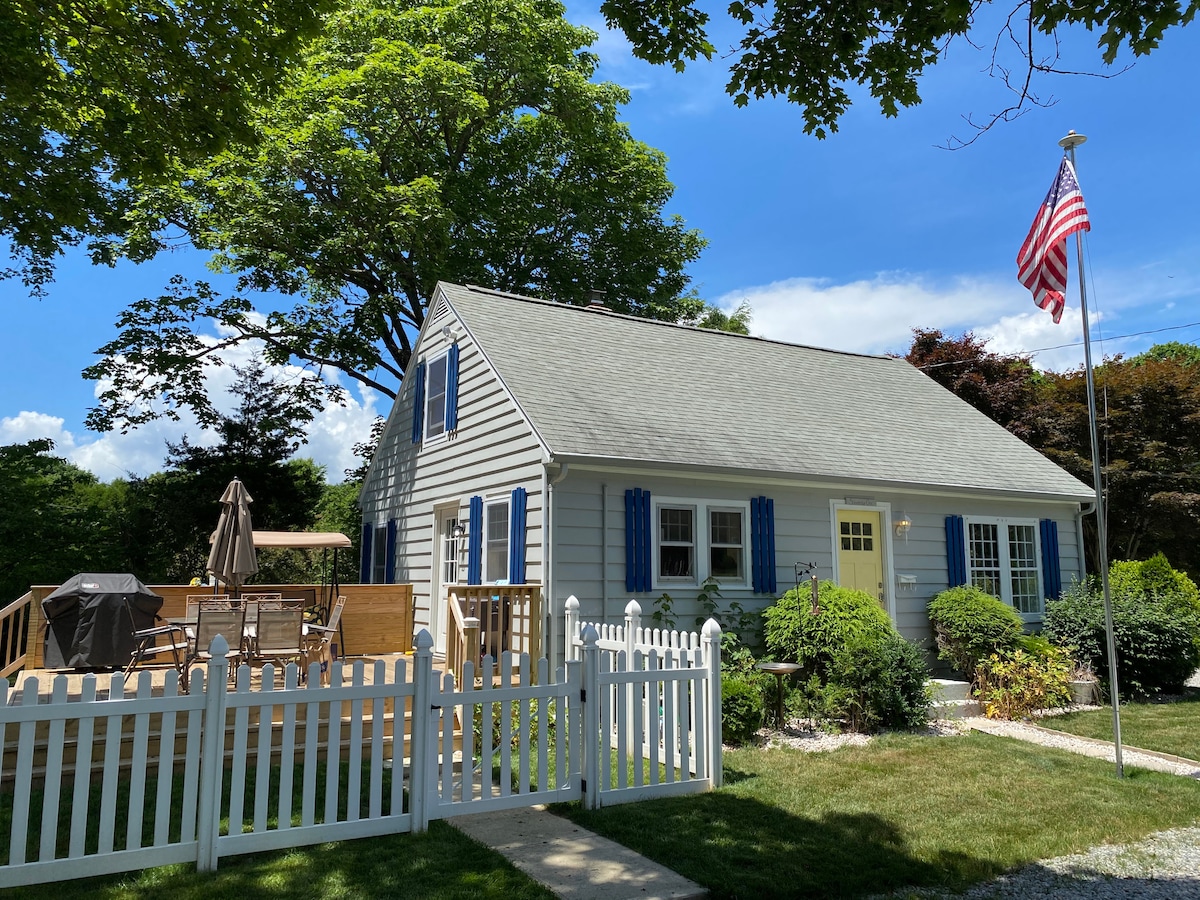 A charming home is framed by a white picket fence and lush greenery. Blue shutters adorn the windows, while an American flag stands proudly. A spacious deck is visible, featuring outdoor furniture and a grill, inviting relaxation and outdoor gatherings.