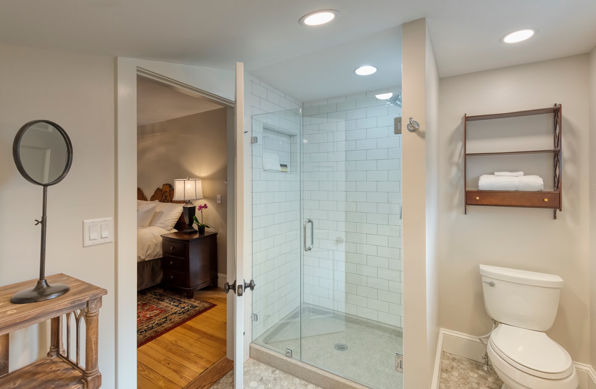 A private bathroom is seen featuring a glass-enclosed shower with white tiles and a modern showerhead. A toilet is positioned nearby, and a wooden shelf holds neatly folded towels. The bathroom connects to a warm sleeping area, visible through the open door.